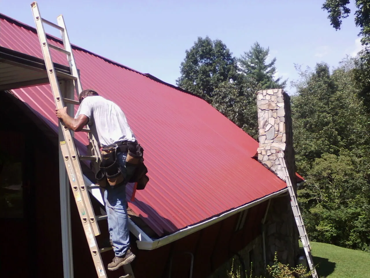 Roofer on a ladder beside a red metal roof and stone chimney — Soffit & Fascia Repair by Roofing Brothers in Dexter City